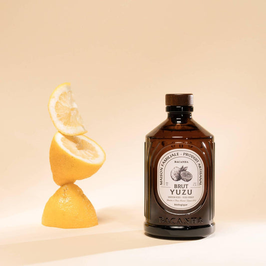 Brown glass bottle of Bacanha Yuzu Syrup on a peach colored background sitting next to a stack of sliced yuzu
